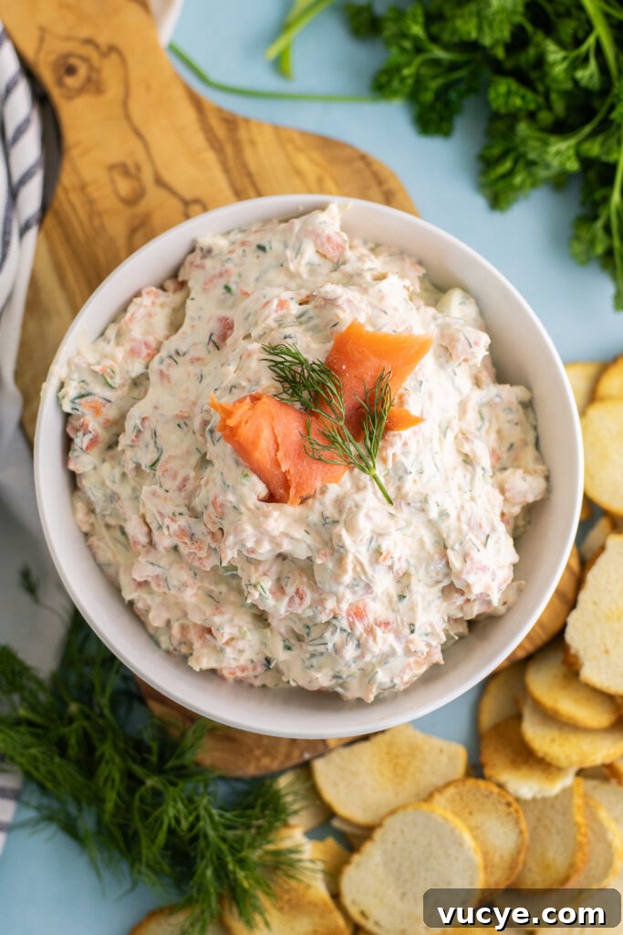 Salmon and Cream Cheese Dip & Spread: Lox-Style Appetizer Recipe 3 Overhead view of smoked salmon dip in a bowl garnished with a slice of smoked salmon and a sprig of fresh dill. The bowl is sitting on a small cutting board next to a pile of bagel chips, fresh dill, and fresh parsley.