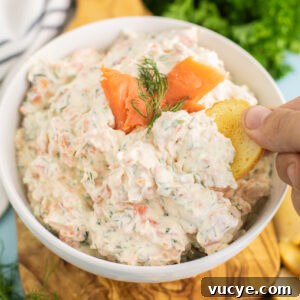 Smoked Salmon Dip in a white ceramic bowl topped with slices of smoked salmon and a sprig of fresh dill with a bagel chip dipping in.