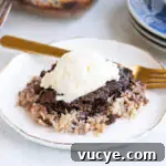 German Chocolate Dump Cake Recipe: Quick Gooey Chocolate Coconut Cake 7 A serving of chocolate and coconut dump cake topped with a scoop of vanilla ice cream on a white plate with a gold fork and blue plates in the background.
