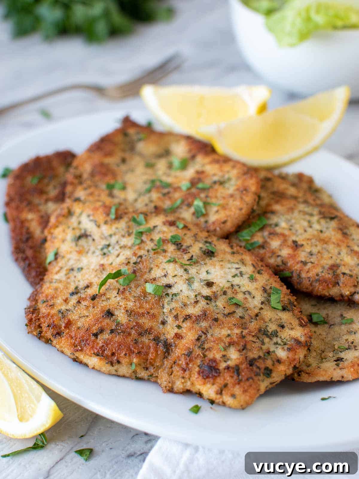 Italian chicken cutlets on white plate with lemon wedges and chopped parsley garnish.