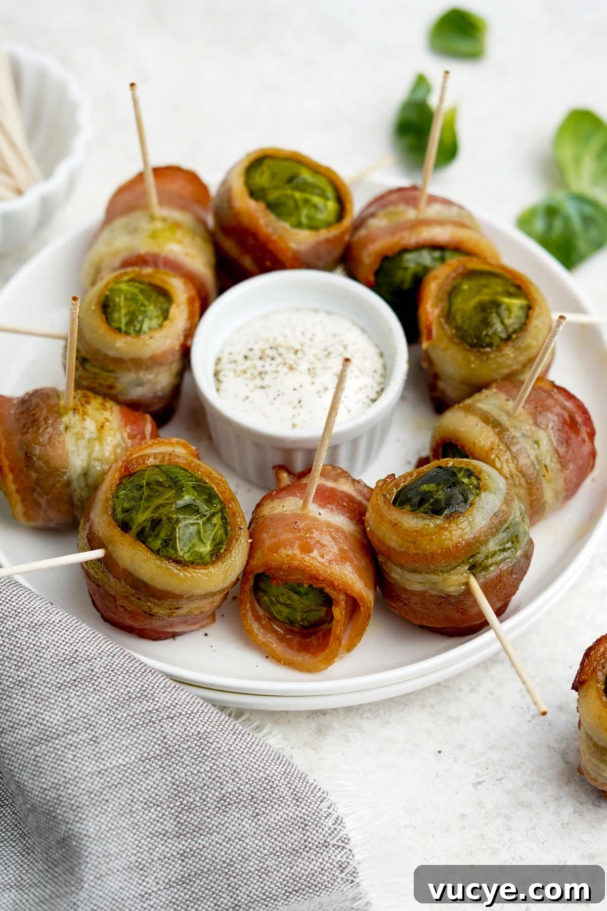 A white plate with bacon wrapped brussels sprouts on it around a small bowl of sauce.