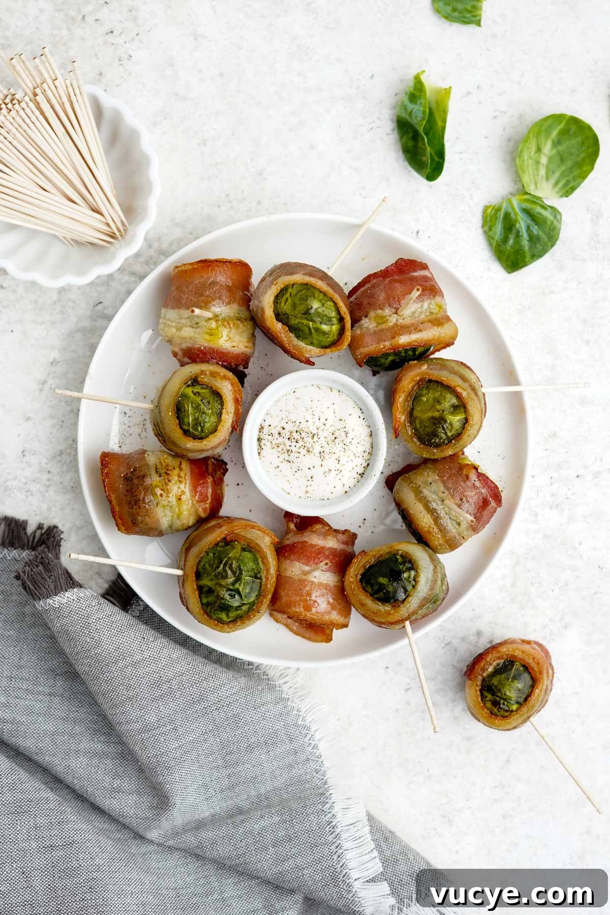  A plate of bacon wrapped brussels sprouts on the table with two on the table next to the plate and a bowl of toothpicks.