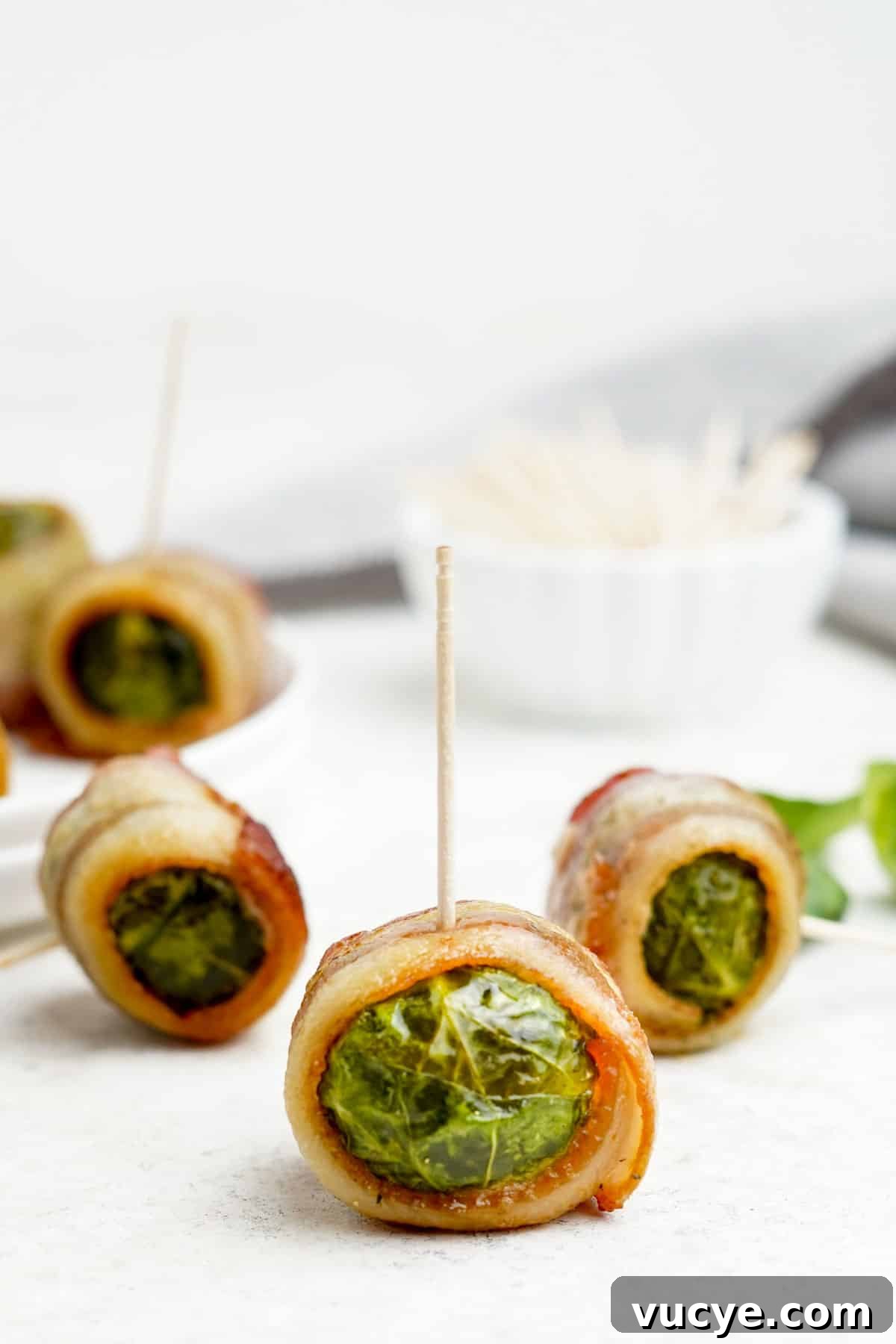A bacon wrapped brussels sprout with a toothpick in it on the table with more of them in the background with a bowl of toothpicks.