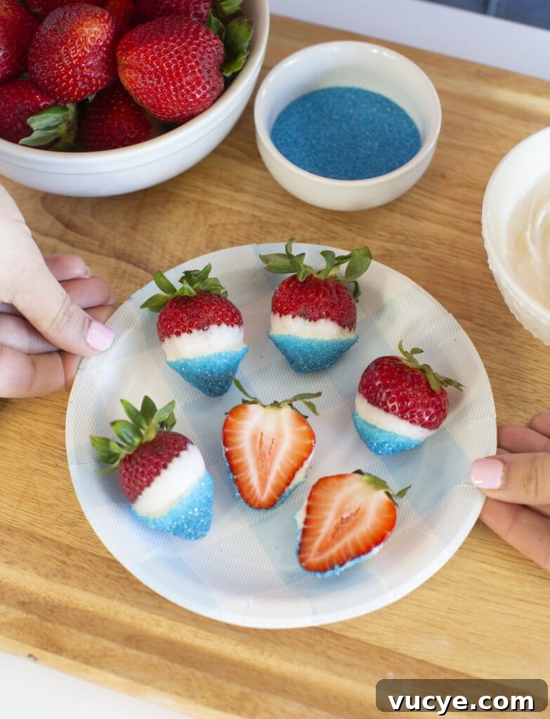 Red, White & Blue Dipped Strawberries for 4th of July 2 A plate of strawberries dipped in white chocolate and blue sugar sits on a wooden surface. Two hands hold the plate. A bowl of plain strawberries, a bowl of blue sugar, and a bowl of white chocolate are nearby.