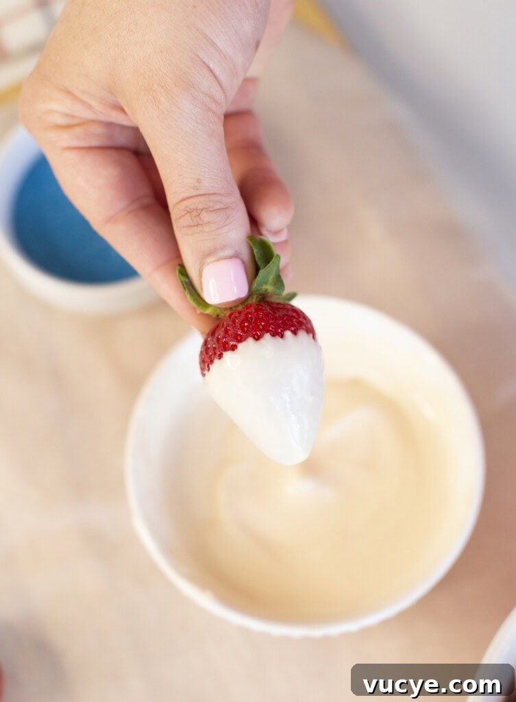 Red, White & Blue Dipped Strawberries for 4th of July 4 A hand with pink nail polish holds a strawberry partially dipped in white chocolate above a white bowl filled with melted white chocolate.
