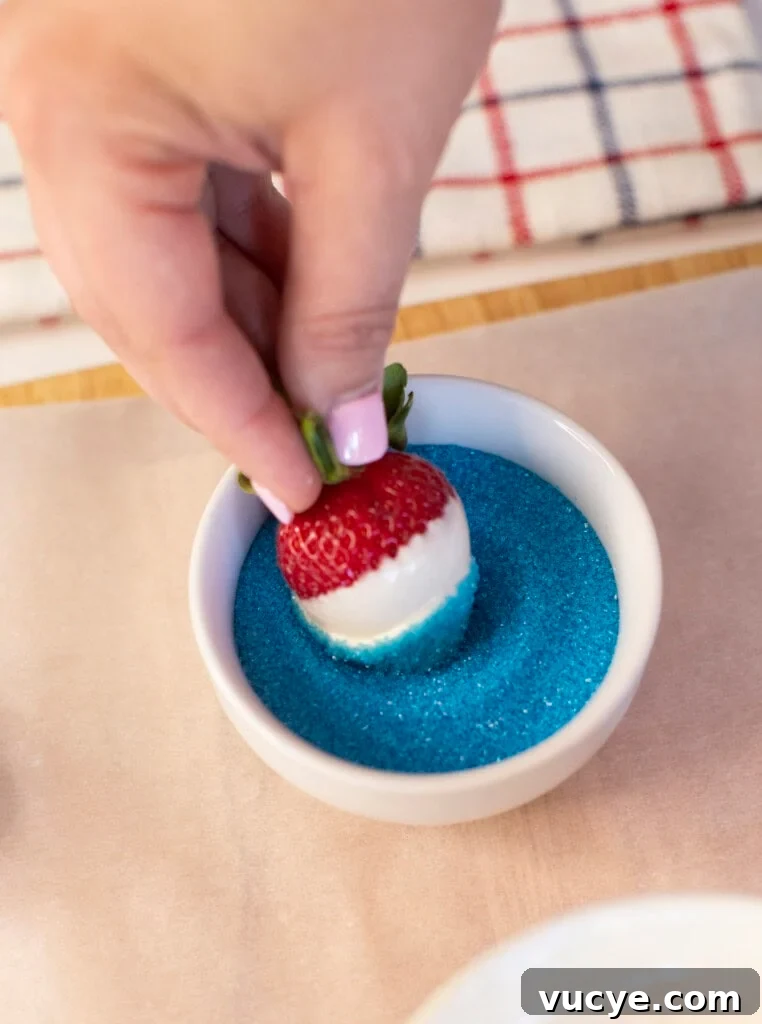 Red, White & Blue Dipped Strawberries for 4th of July 5 A hand dips a white chocolate-covered strawberry into a bowl of blue sugar, creating a red, white, and blue treat. A striped cloth is visible in the background.