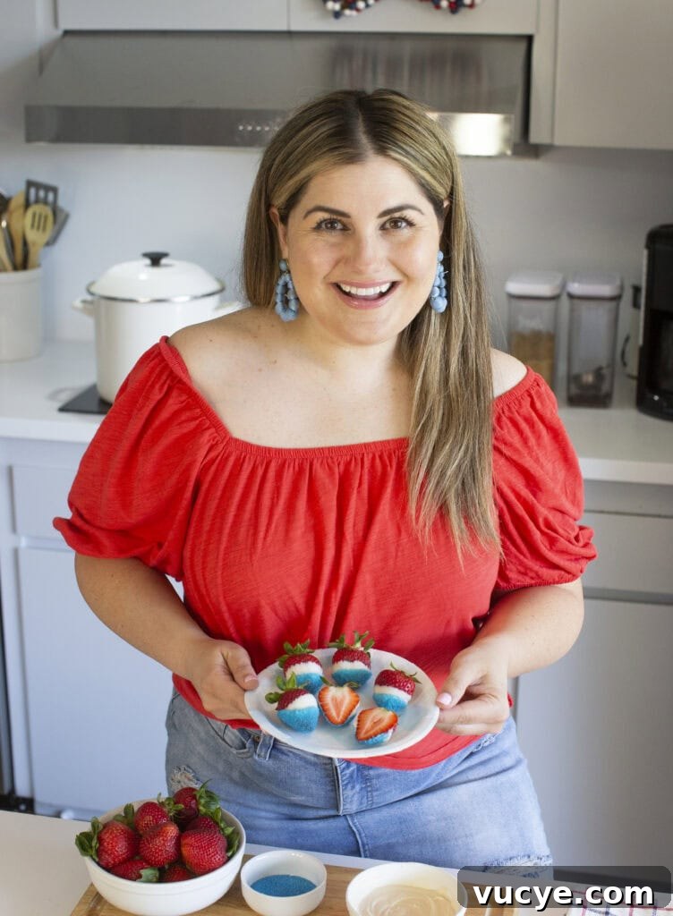Red, White & Blue Dipped Strawberries for 4th of July 6 A woman in a red off-the-shoulder top holds a plate of decorated strawberries in a modern kitchen. Bowls of strawberries, blue sprinkles, and white chocolate are on the counter in front of her.