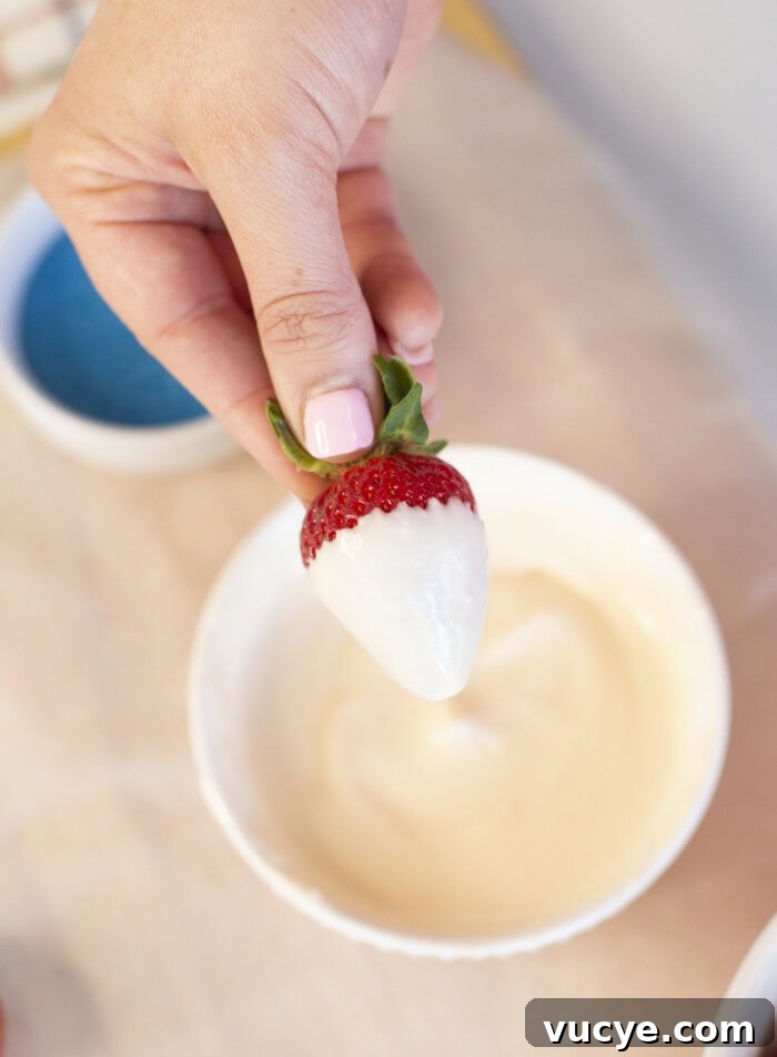 Red, White & Blue Dipped Strawberries for 4th of July 8 A hand with pink nail polish holds a strawberry partially dipped in white chocolate above a white bowl filled with melted white chocolate.