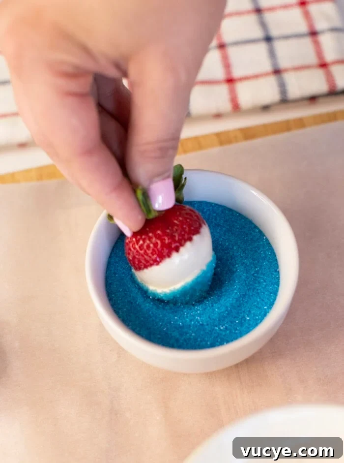Red, White & Blue Dipped Strawberries for 4th of July 9 A hand dips a white chocolate-covered strawberry into a bowl of blue sugar, creating a red, white, and blue treat. A striped cloth is visible in the background.