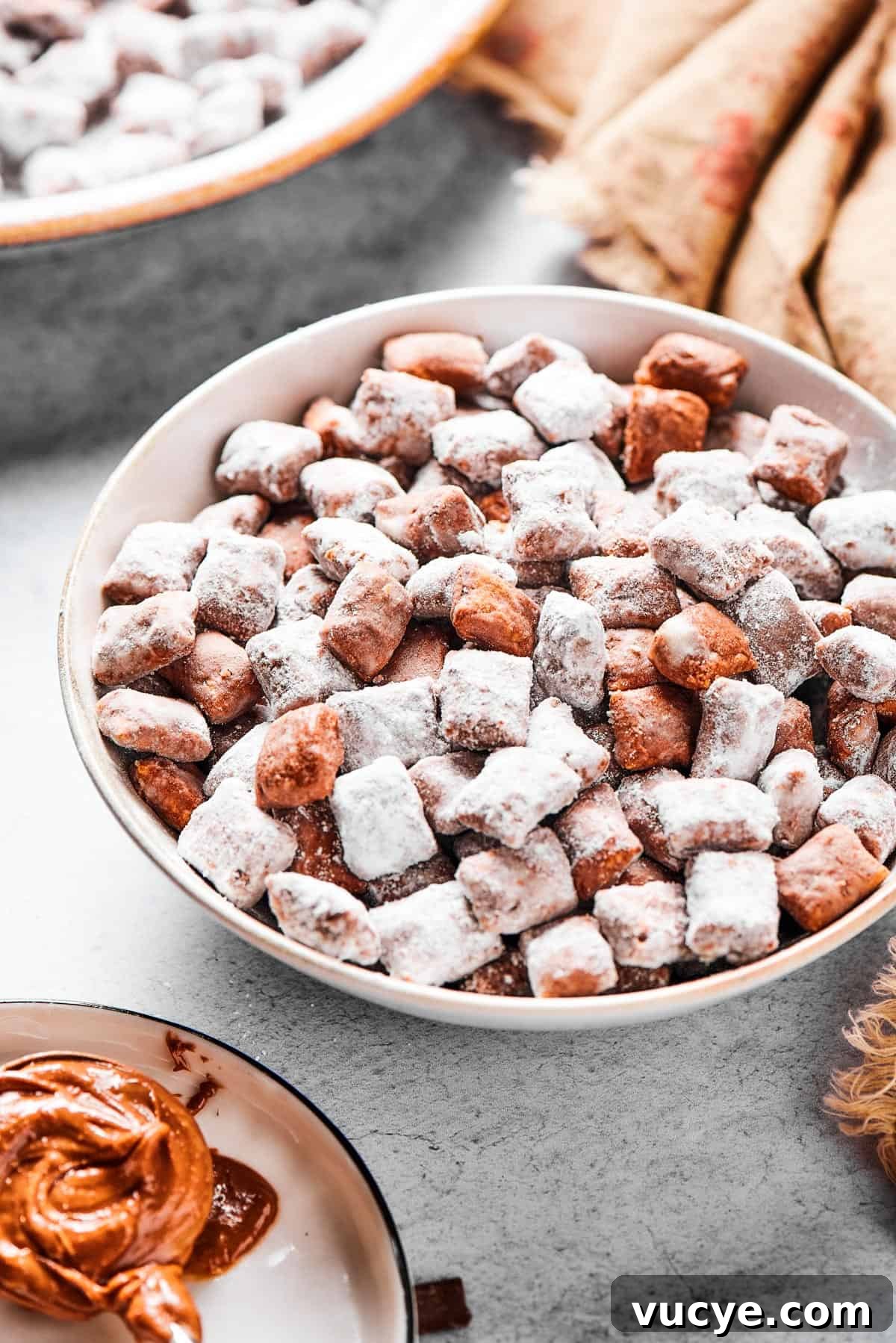 Who’s up for Muddy Buddies? Smooth chocolate, salty peanut butter, and a light dusting of powdered sugar make this crispy Chex cereal snack a true favorite! A shallow bowl filled with candy-coated cereal, near a beige napkin. Peanut butter and chocolate chunks are nearby on the table.