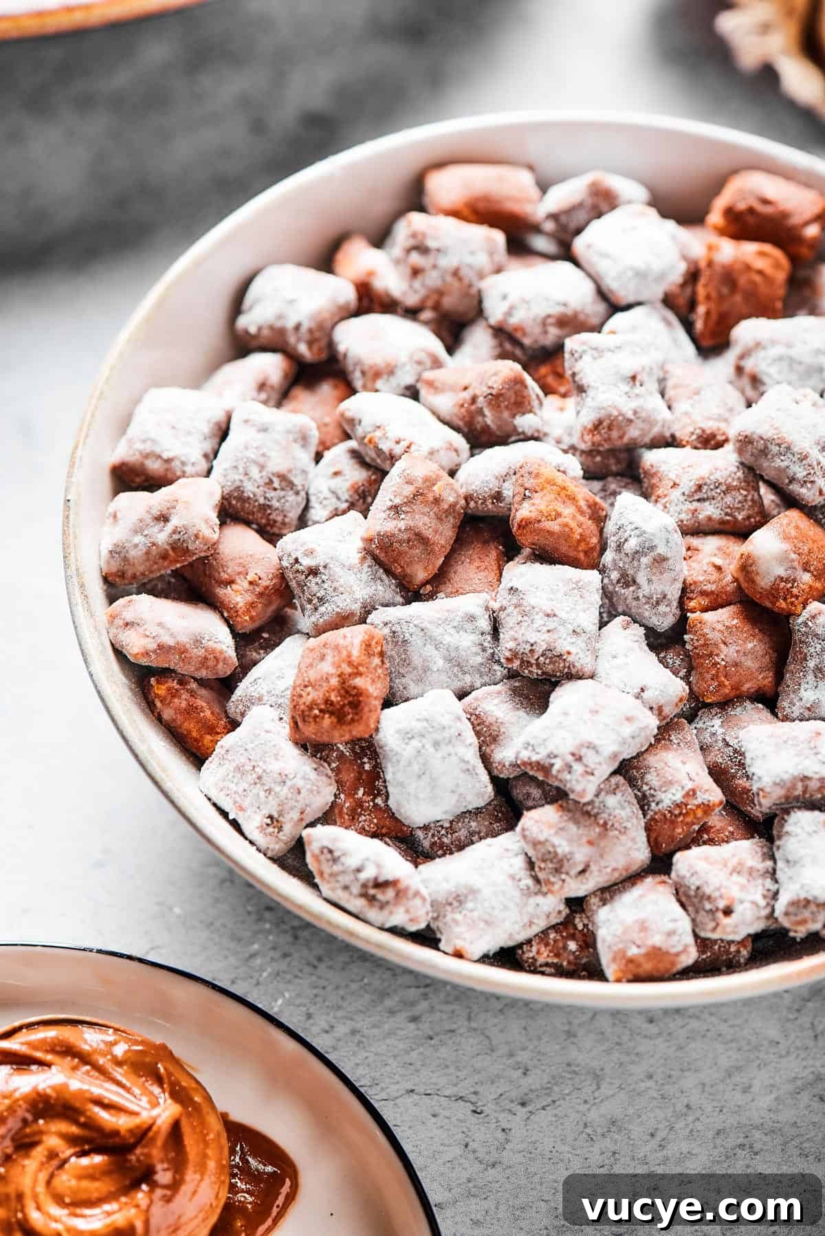Crunchy Peanut Butter Muddy Buddies Recipe 8 Close-up shot of puppy chow in a white bowl, to show the texture. Some pieces are fully coated in powdered sugar, while others are not.