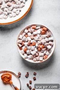 Crunchy Peanut Butter Muddy Buddies Recipe 7 Muddy buddies in a white bowl, with a spoonful of peanut butter and a larger serving dish of the snack nearby.