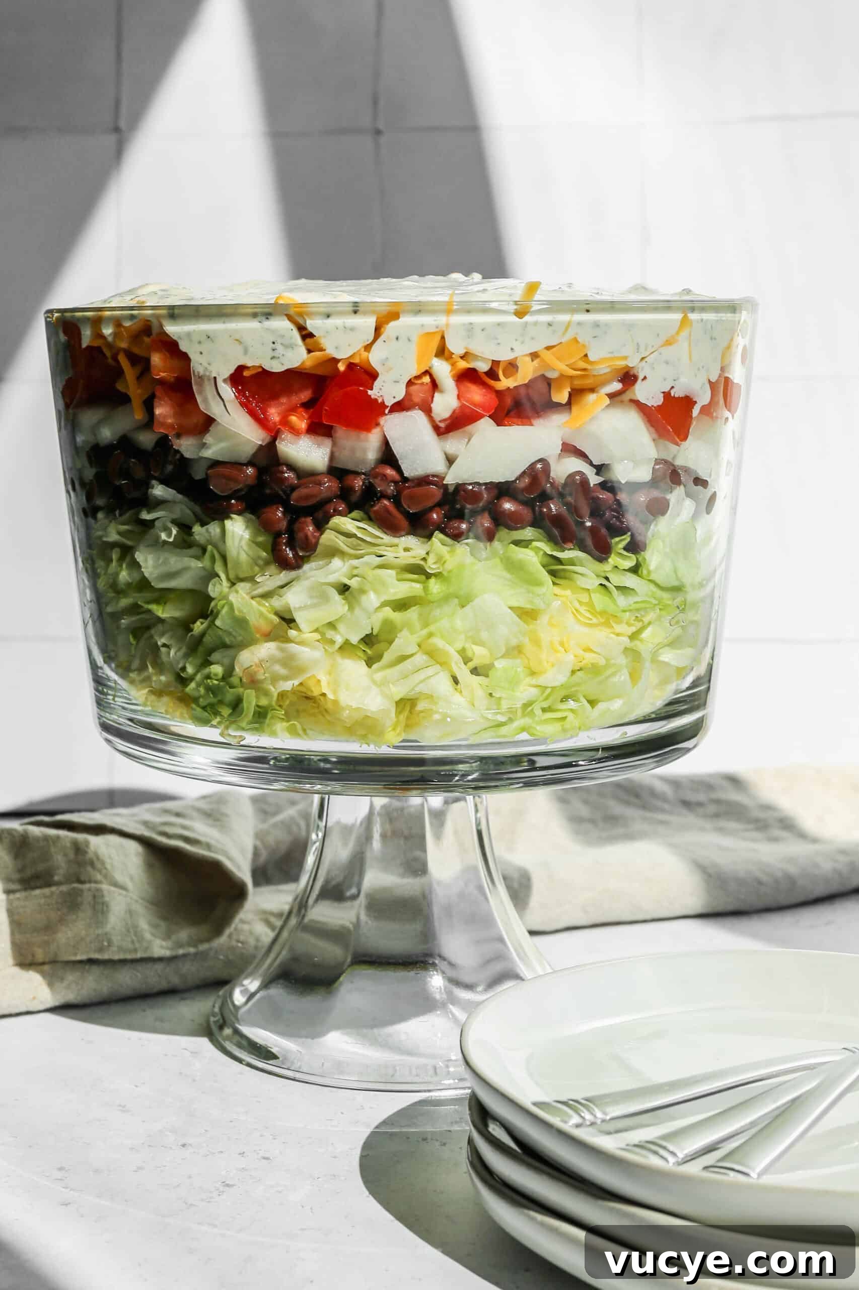 a glass trifle dish of chopped wedge layer salad next to a stack of white plates.