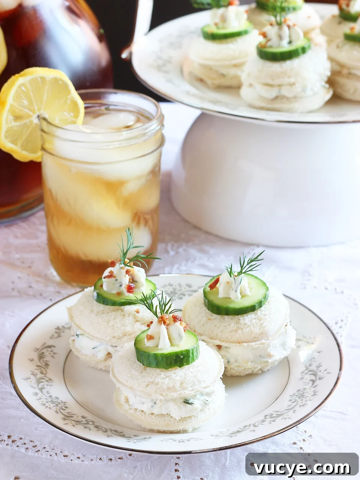 Cucumber Tea Sandwich Recipe for Light, Refreshing Tea Parties 2 Three cucumber tea sandwiches garnished with a dollop of cream cheese, cucumber slice, fresh dill and bacon bits. Platter of sandwiches and a glass of iced tea in the background.