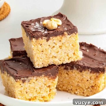 A stack of four chocolate peanut butter Rice Krispie treats on a plate, each with a glossy chocolate top and lightly salted chopped peanuts for garnish.