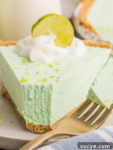 Close-up of a slice of key lime Jello pie with a bite on a fork beside it. The pie is topped with whipped cream, lime zest, and a lime slice, showing its airy and creamy whipped texture.