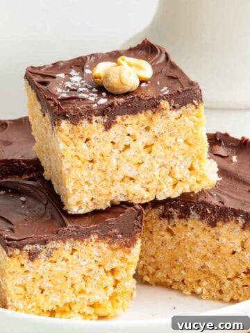 A stack of four chocolate peanut butter Rice Krispie treats on a plate, each with a glossy chocolate top and lightly salted chopped peanuts for garnish.