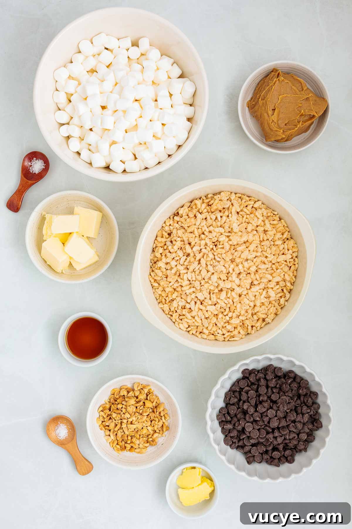 Flat lay of measured ingredients in bowls for making Rice Krispie treats, including mini marshmallows, butter, peanut butter, vanilla, chopped peanuts, chocolate chips, and crispy rice cereal.