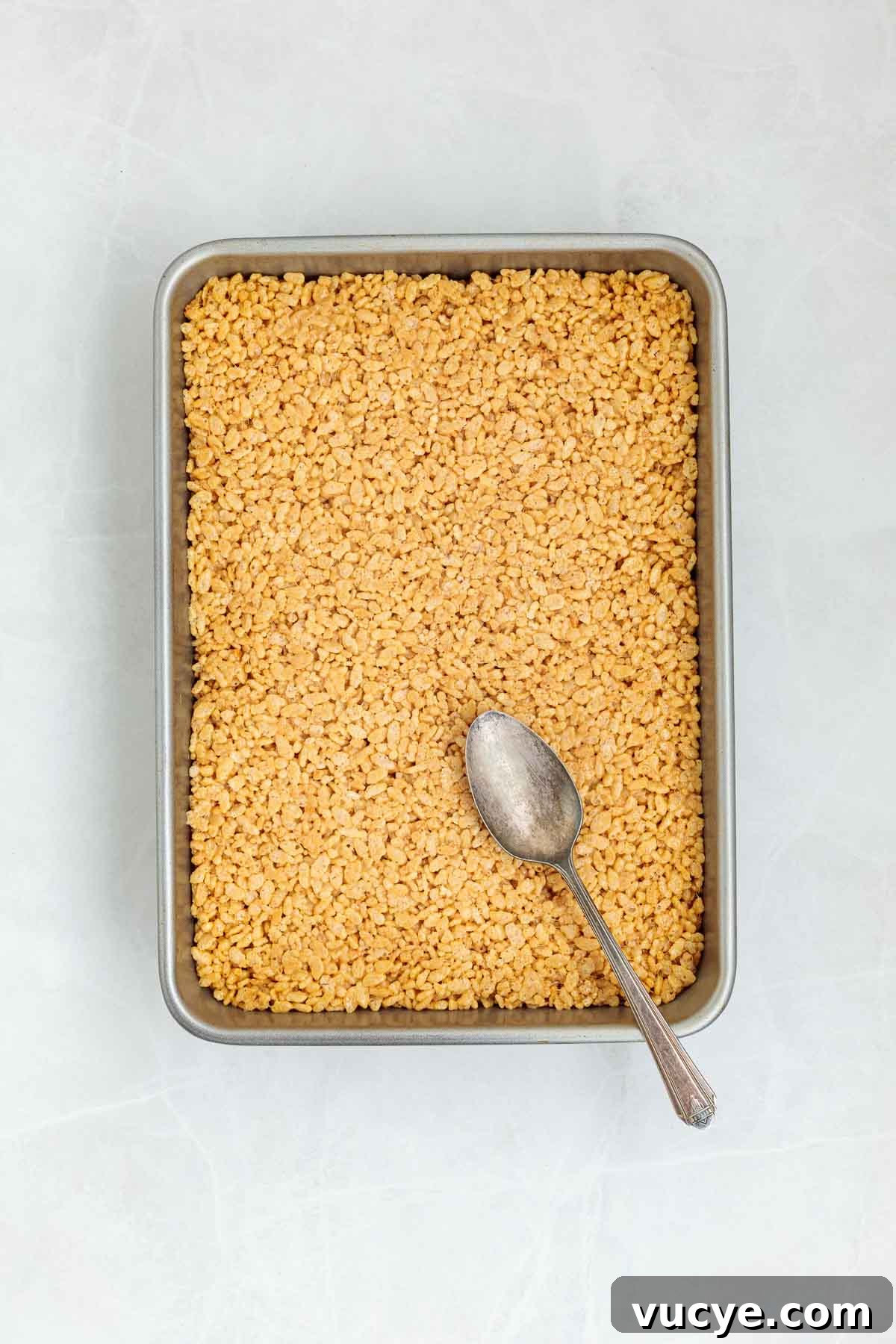 Rice Krispie mixture pressed evenly into a metal baking pan with the back of a spoon resting on top, ready to be topped or cooled.