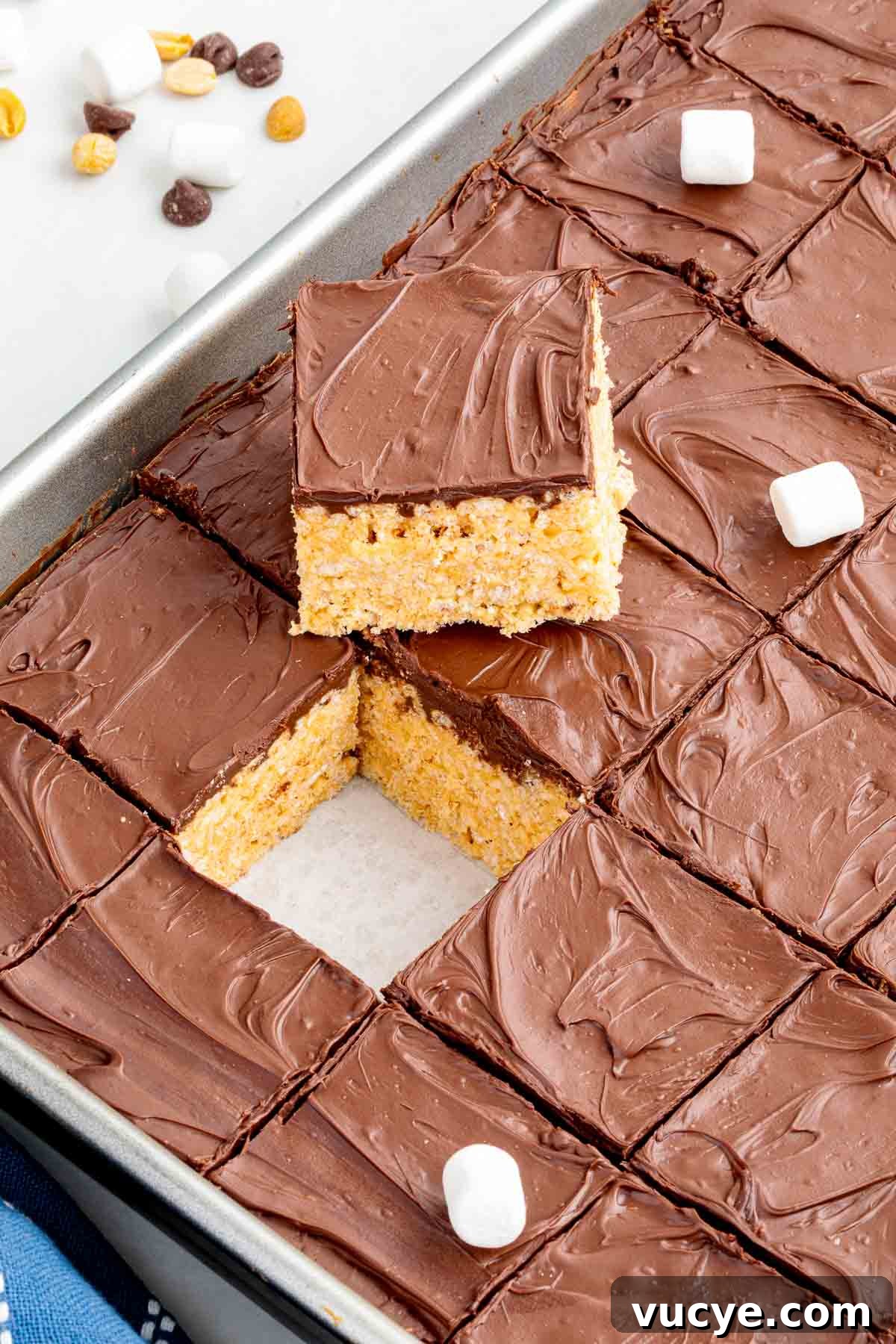 Tray of sliced chocolate-topped Rice Krispie treats with one square lifted out, revealing the crispy peanut butter base underneath.