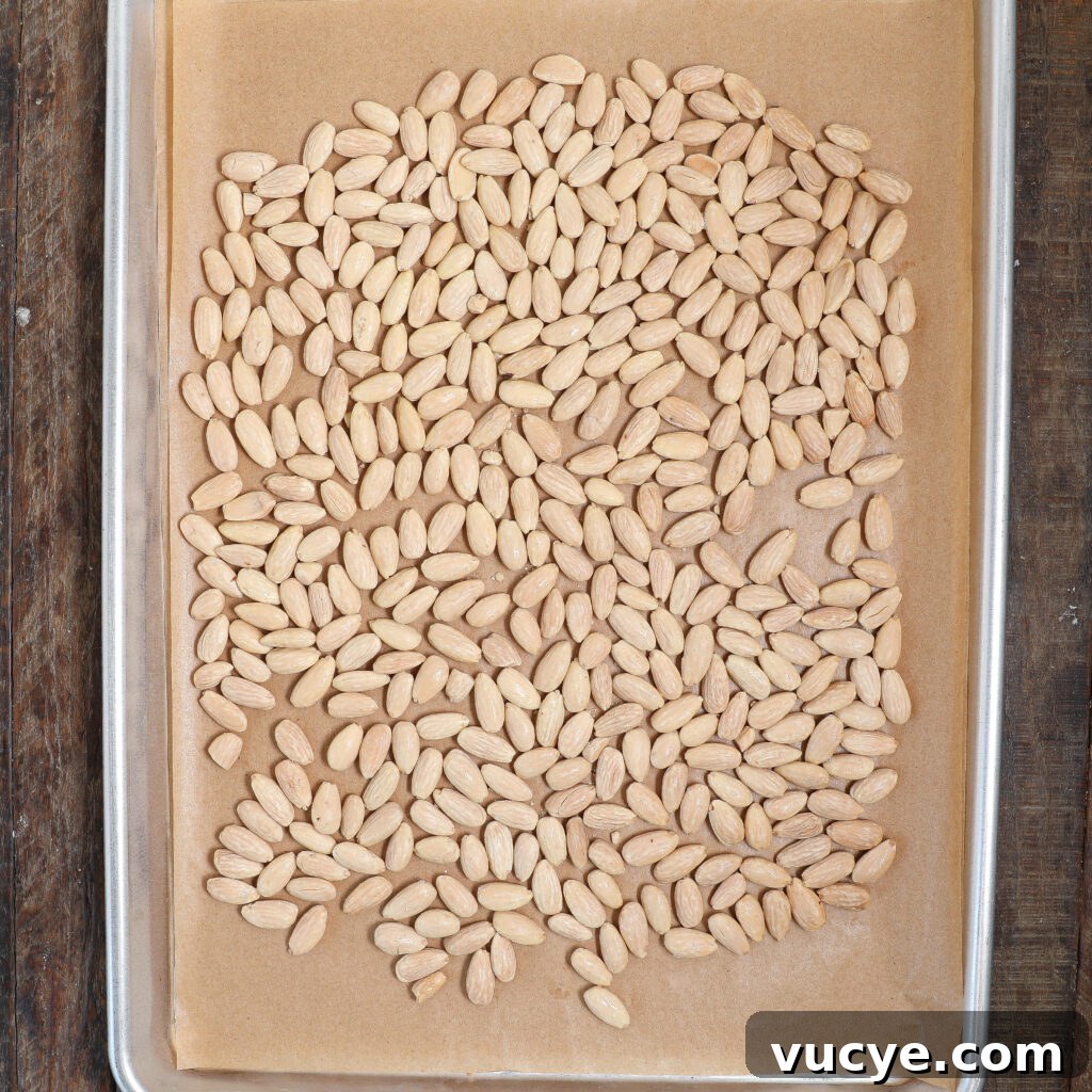 A baking sheet lined with parchment paper holds a single layer of blanched almonds, spread out evenly and ready for roasting—perfect for making Chocolate Bark with Honey Glazed Almonds on a wooden surface.