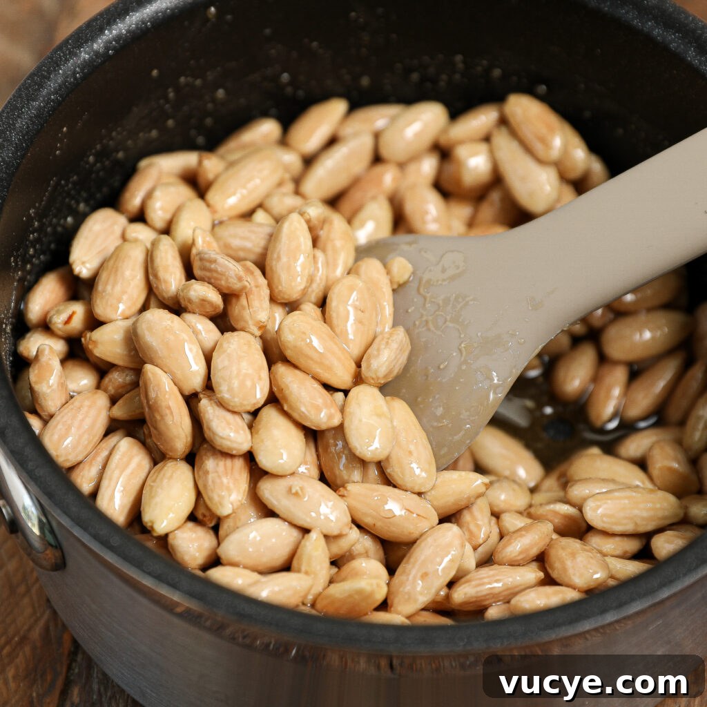 A close-up of a pot filled with whole blanched almonds being stirred with a beige spatula. The glossy almonds, likely coated in syrup or oil, are perfect for making Chocolate Bark with Honey Glazed Almonds.