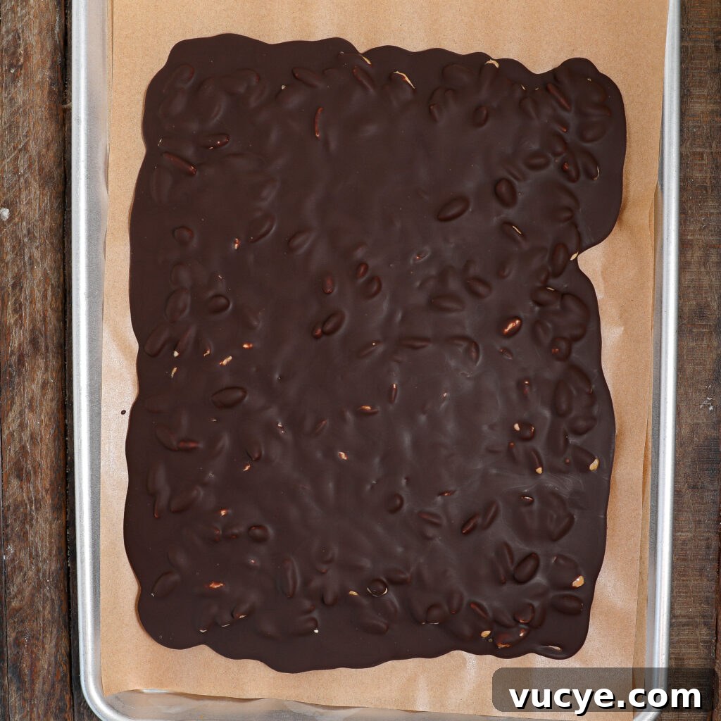 A sheet pan lined with parchment paper holds a large slab of chocolate bark with honey glazed almonds and visible nuts mixed in, resting on a wooden surface.