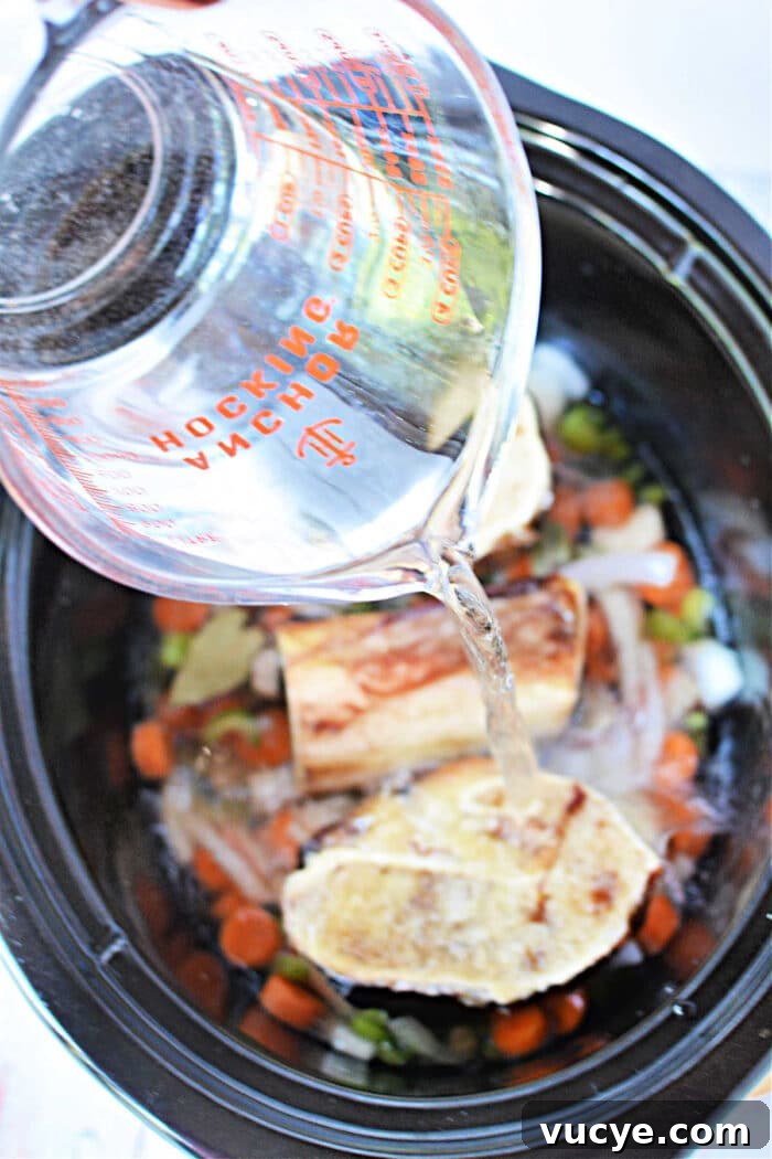 Pouring water into a slow cooker, creating a rich bone broth with vegetables and bones.