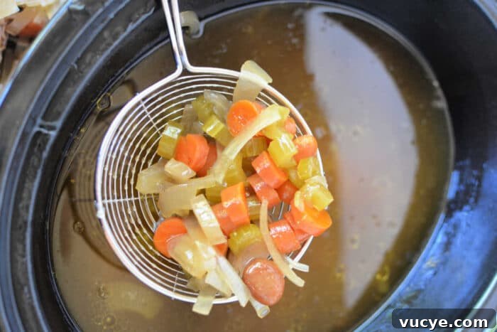 A wire ladle lifts diced carrots, celery, and onions above a pot of simmering bone broth, creating a fragrant medley perfect for your slow cooker.