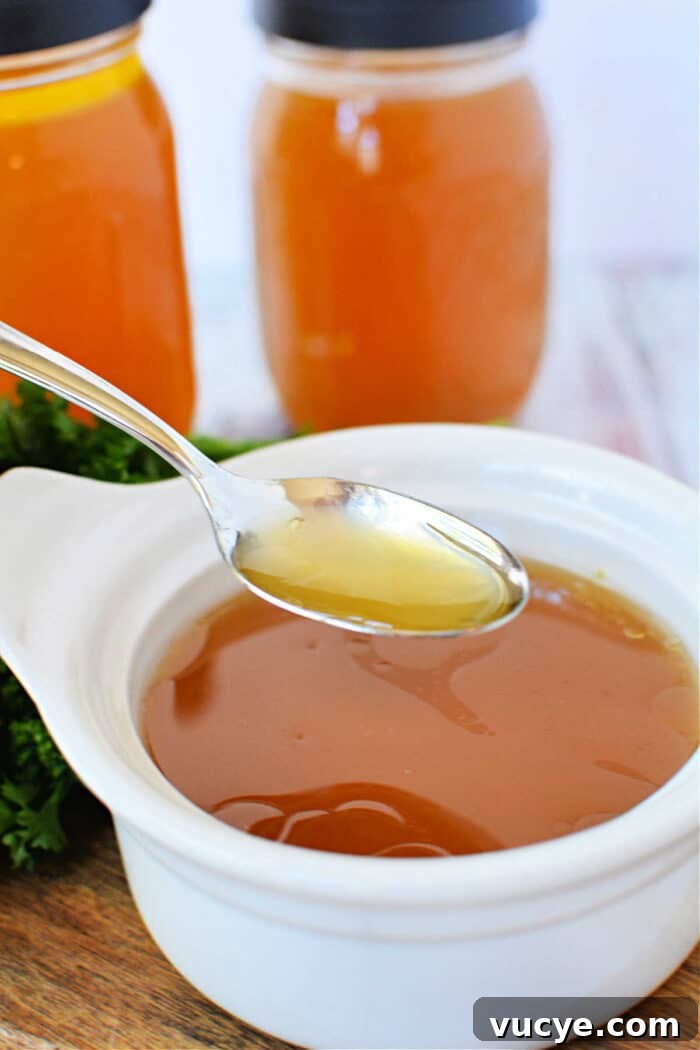 A spoon hovers over a white bowl, filled with homemade bone broth made in a slow cooker, with two jars in the background.