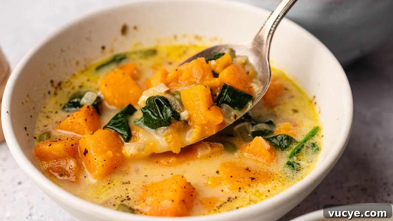 A bowl of creamy soup containing chunks of orange squash, leafy spinach, and visible seasoning, with a spoon lifting a portion.