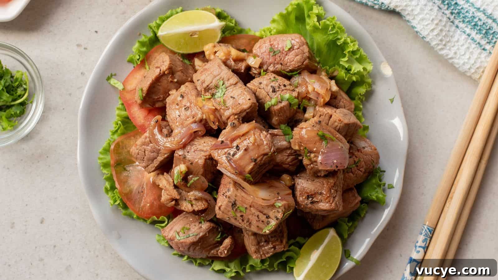 A plate of stir-fried beef cubes with onions, garnished with cilantro, served on lettuce with tomato slices and lime wedges. Chopsticks rest beside the plate.