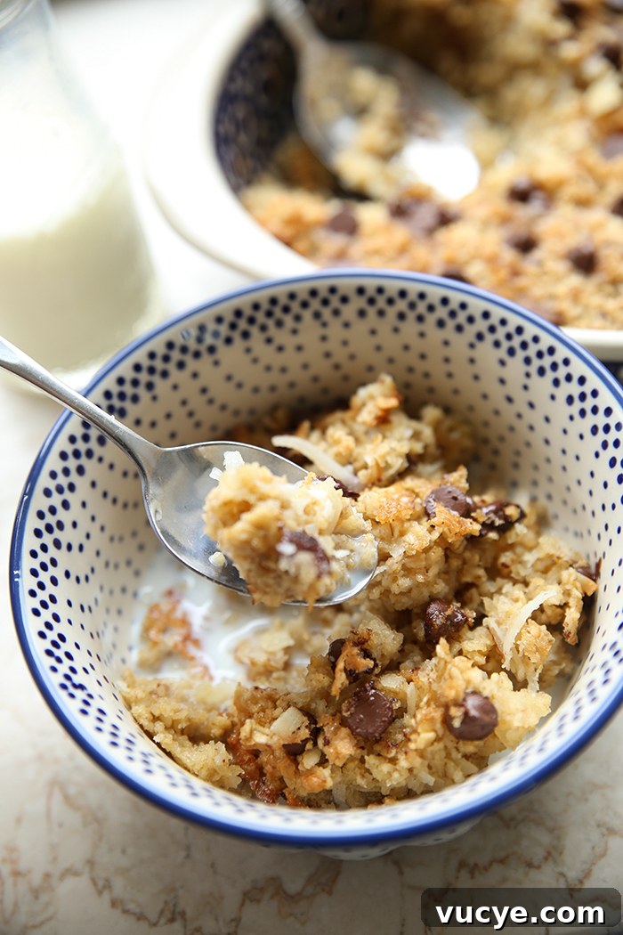 A white bowl with blue dots with a serving of almond joy baked oatmeal with milk. A spoon with oatmeal on it is placed on the rim of the bowl and on top of the oatmeal. Faded in the background is a glass of milk and a baking dish of oatmeal with a serving scooped out and a spoon set in the empty space.