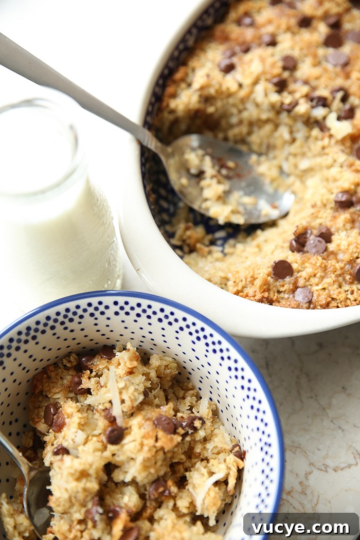 Almond Joy baked oatmeal in a Polish pottery bowl with a white background and blue dots with a spoon inserted diagonally from the left. A glass of milk is above the bowl on the left. Diagonally placed from the upper right corner is an oval baking dish of almond joy oatmeal with a scoop taken out and a spoon in its place lying diagonally from the left. 