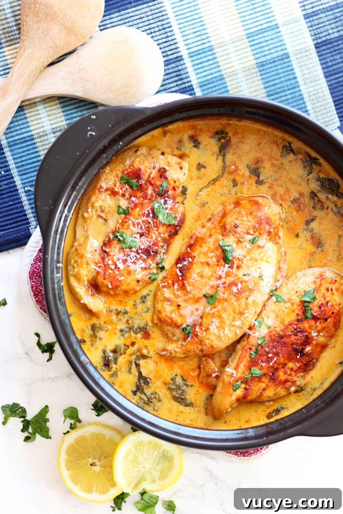Black pan containing 3 Lemon Butter Chicken Breasts, crossed spoons and lemon slices on table.