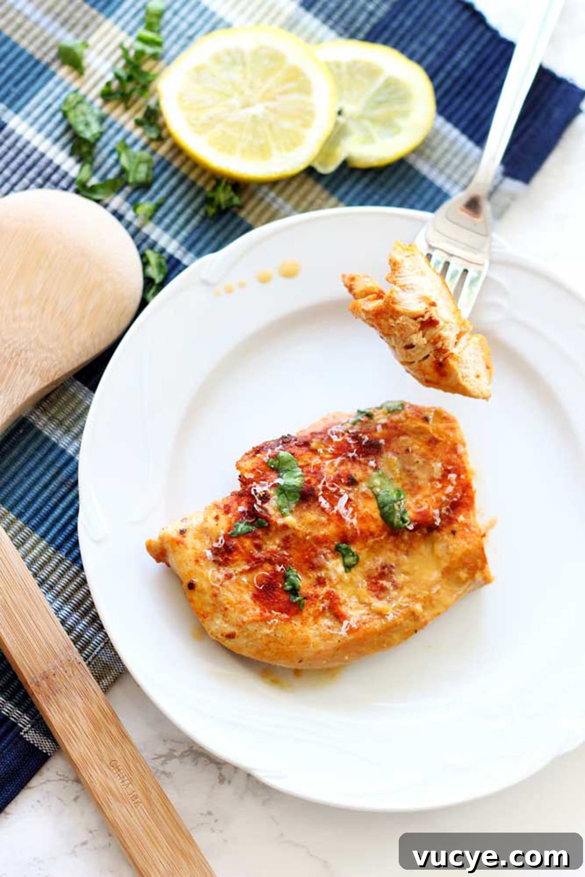 White plate containing a breast of lemon butter chicken, fork in a piece of chicken.