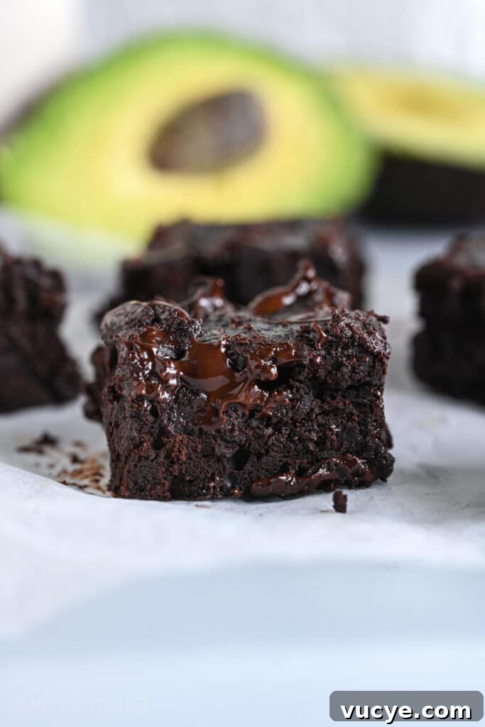 Close-up of a fudgy chocolate avocado brownie with ripe avocados shown in the background.