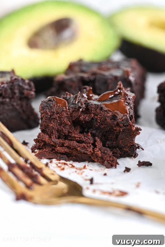 Fudgy avocado brownie after tasting the texture with a golden fork – rich crumbles of chocolate brownie are shown over a white background with rice halved avocados in the background