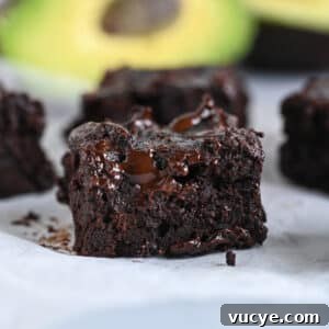 Close-up of a fudgy chocolate avocado brownie with ripe avocados shown in the background.