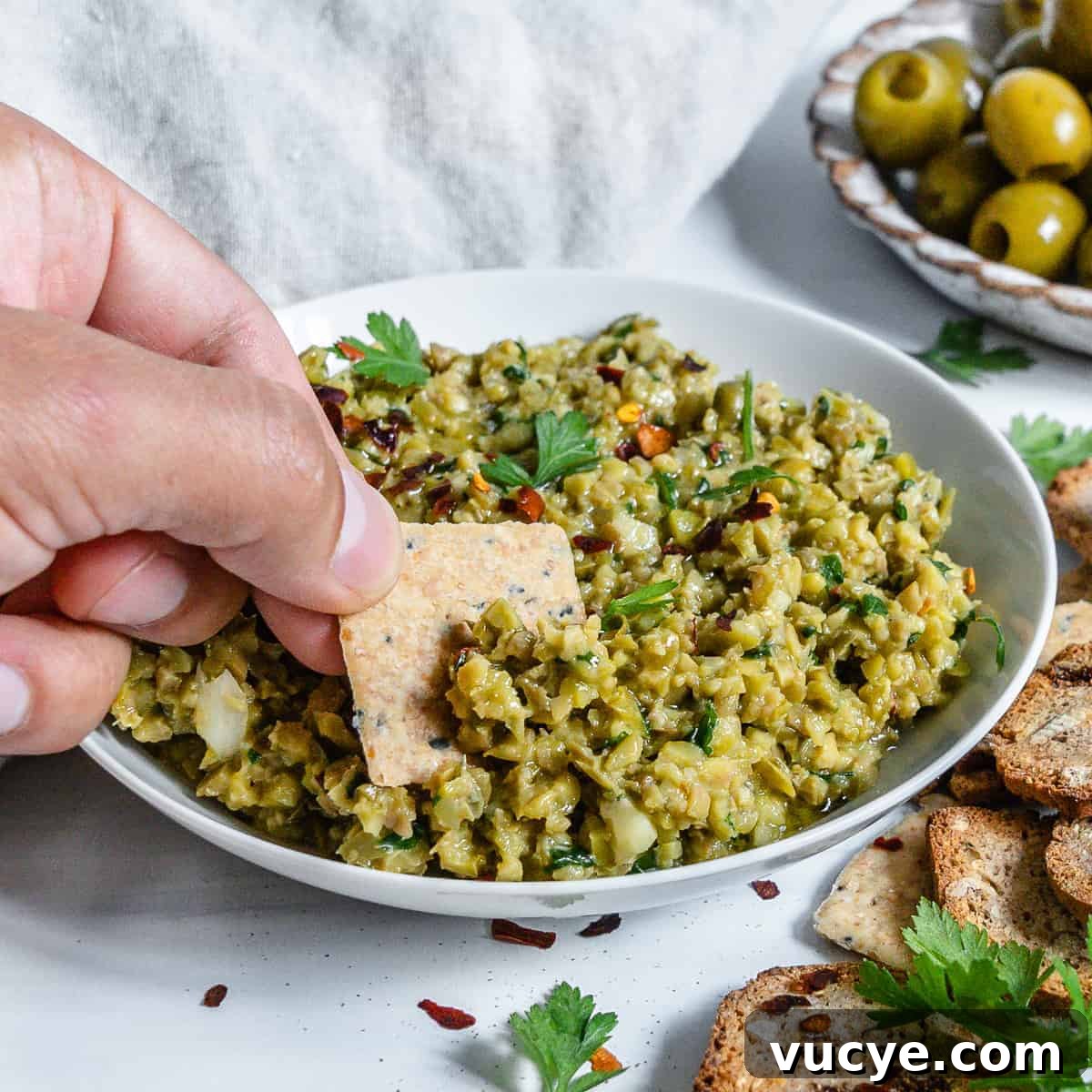 Completed Green Olive Tapenade in a white bowl with a cracker being dipped in the bowl and ingredients in the white background.
