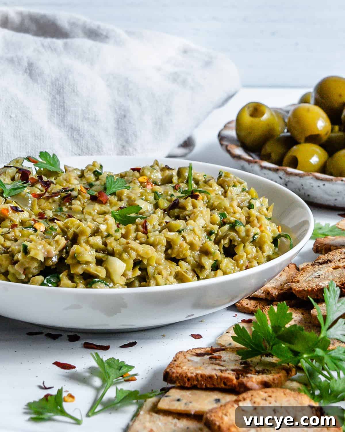 Completed Green Olive Tapenade in a white bowl with a cracker being dipped in the bowl and ingredients in the white background.