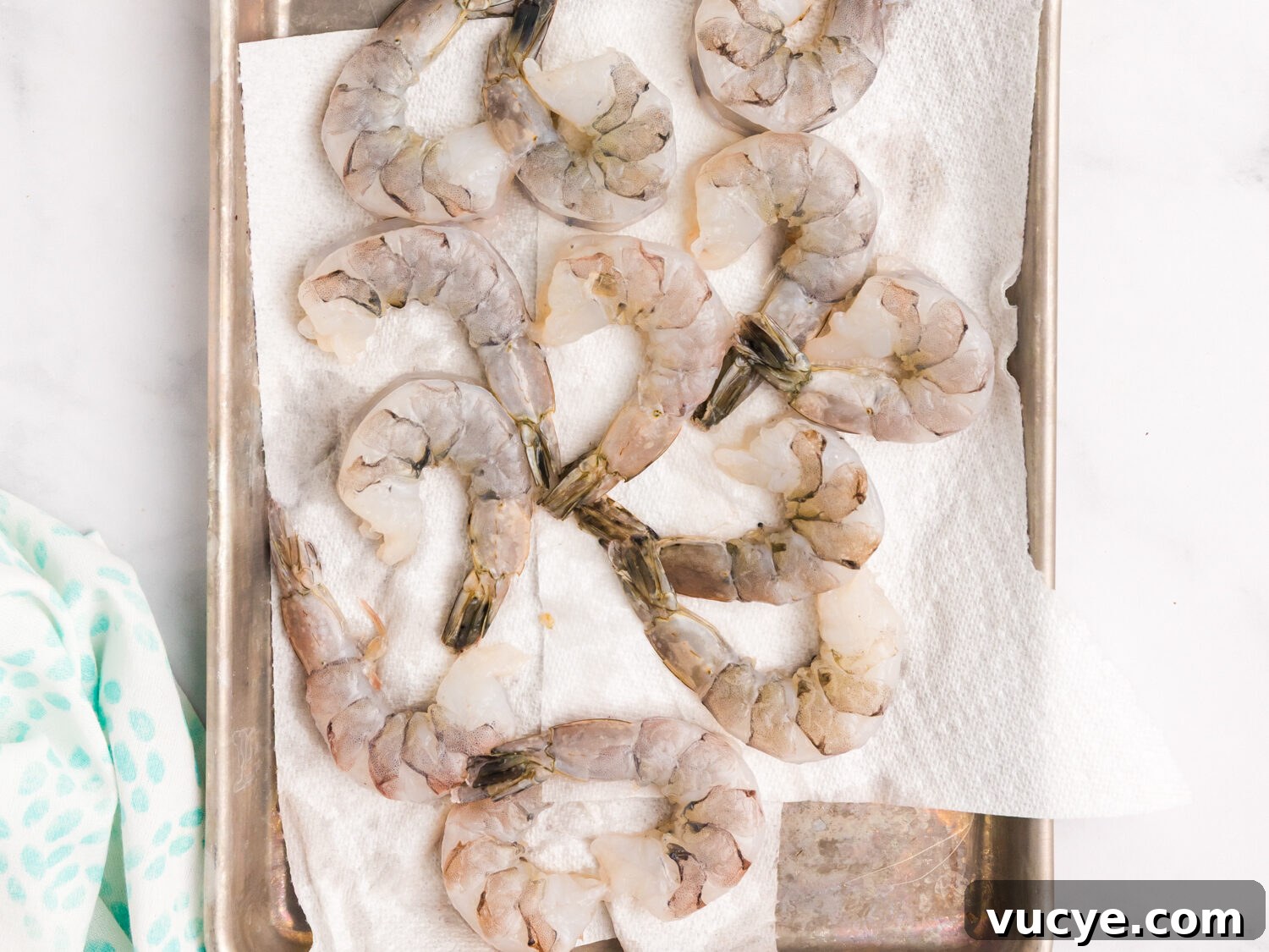 Raw, peeled shrimp laid out on a metal tray lined with paper towels, surrounded by lemon wedges, olive oil, seasoning mix, and salt and pepper shakers.