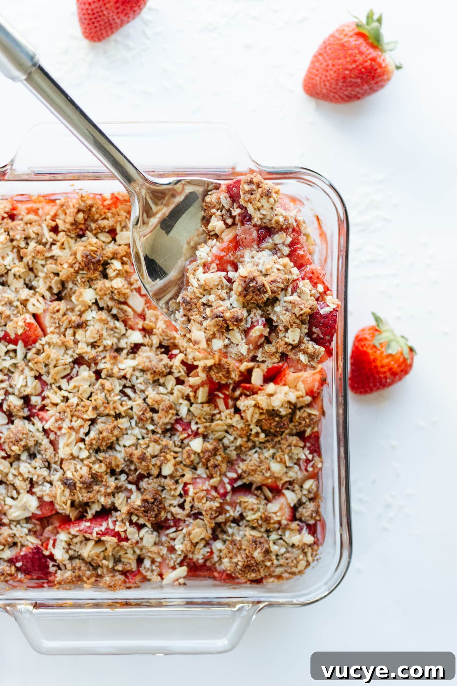 Must-Try Strawberry Recipes for Every Season 13 Square glass dish of strawberry coconut crisp with large metal serving spoon lifting out a portion.