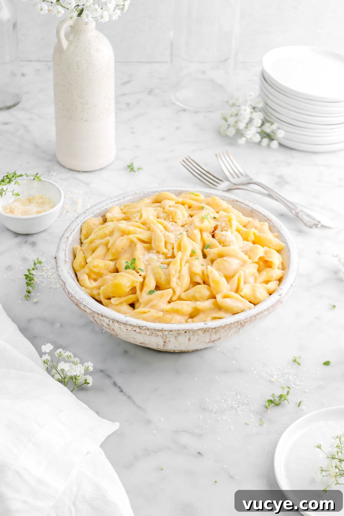 Creamy Four-Cheese Macaroni and Cheese Recipe 5 angled shot of macaroni and cheese in antique bowl with forks, vase of flowers, and bowl of grated parmesan behind