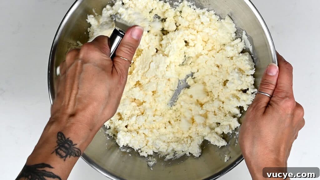 Tiropita: Authentic Greek Cheese Pie Recipe You Can Make at Home 4 Two hands mix crumbly, pale dough in a large metal bowl—one holds a pastry blender, the other steadies the bowl.