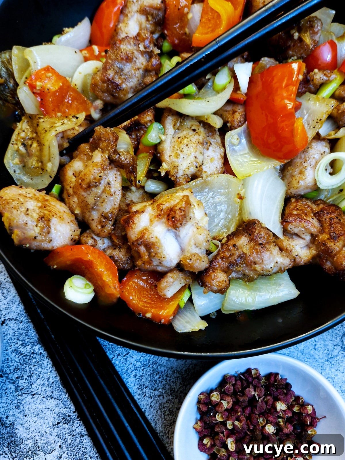 Crispy Salt and Pepper Chicken Recipe for Dinner 15 Close up of pieces of salt and pepper chicken with onions and red peppers in a black serving dish with chopsticks. There is a bowl of Sichuan peppercons in the foreground.