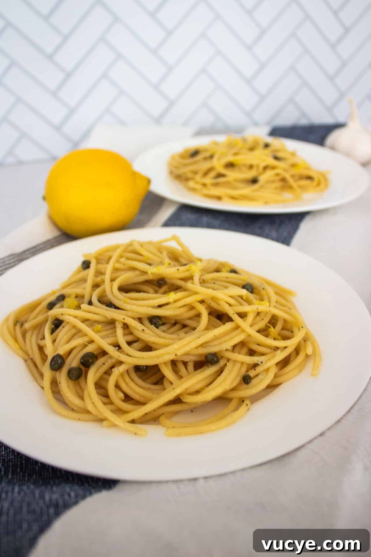 Zesty Lemon Caper Pasta with Garlic and Parmesan 2 Two plates with spaghetti, capers, and lemon zest with a lemon between them and a garlic clove behind.