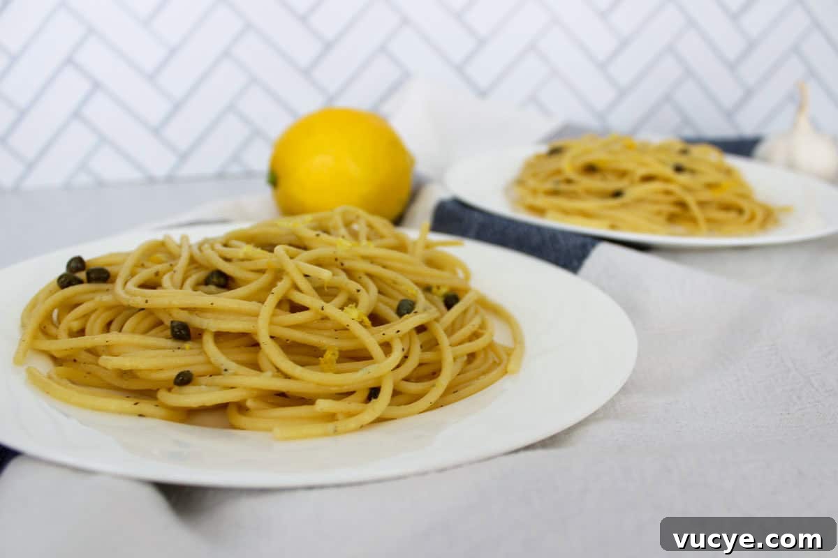 Zesty Lemon Caper Pasta with Garlic and Parmesan 7 Two plates with spaghetti, capers, and lemon zest with a lemon between them and a garlic clove behind.