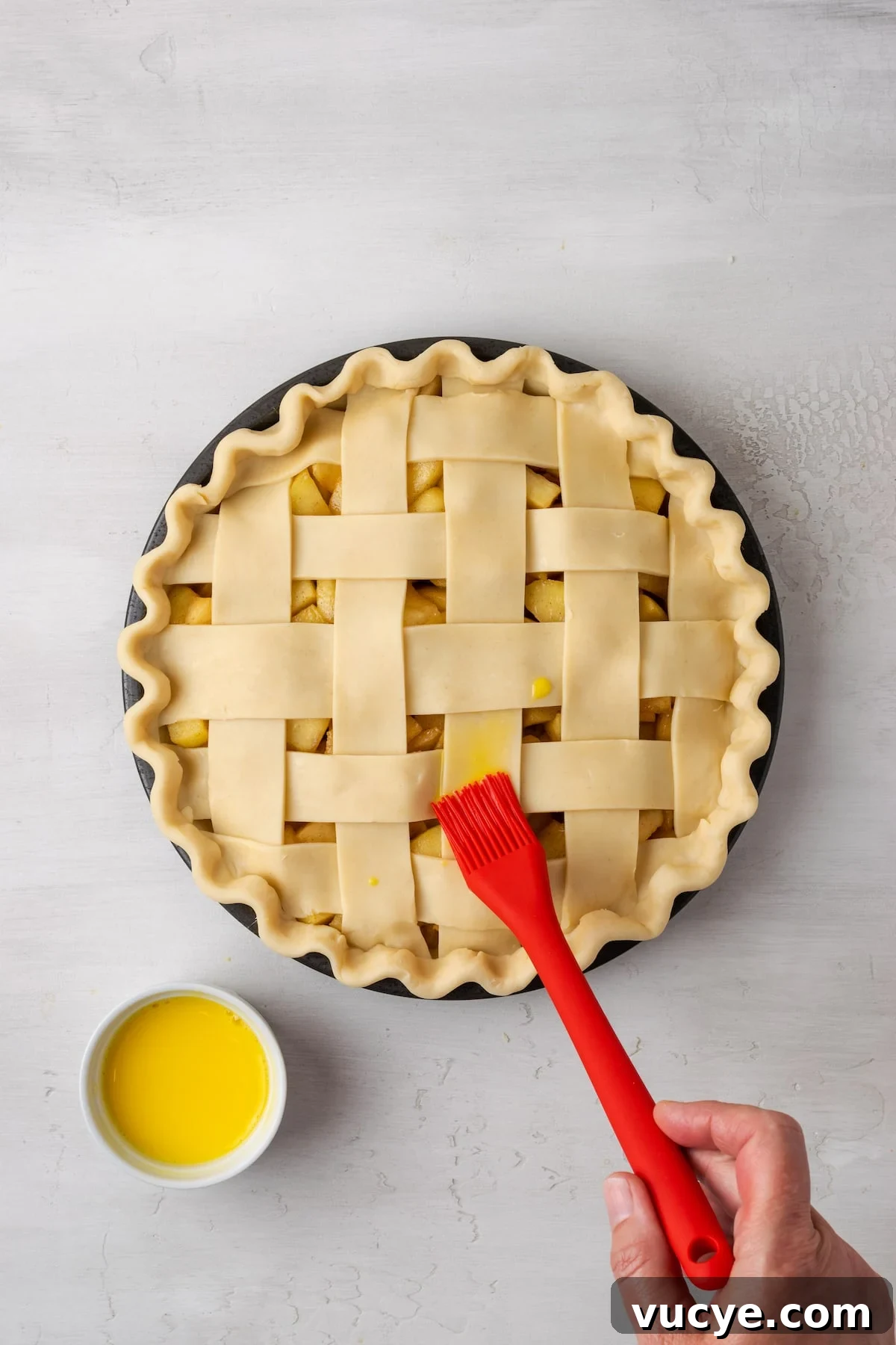 Add an egg wash. A red rubber brush is used to brush egg wash over an unbaked apple pie with a lattice top next to a small bowl of egg wash.