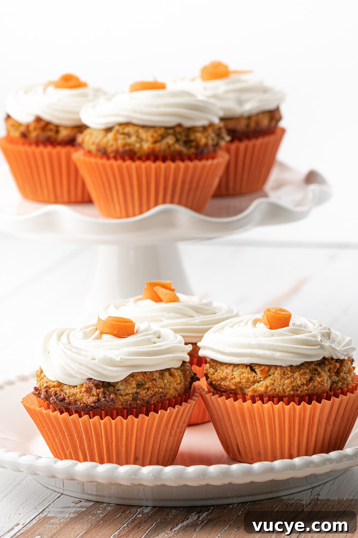 Low-Carb Carrot Cake Muffins with Cream Cheese Frosting 4 White plate with three muffins in the foreground and a stack of carrot cake muffins on a white ruffled cake stand in the back
